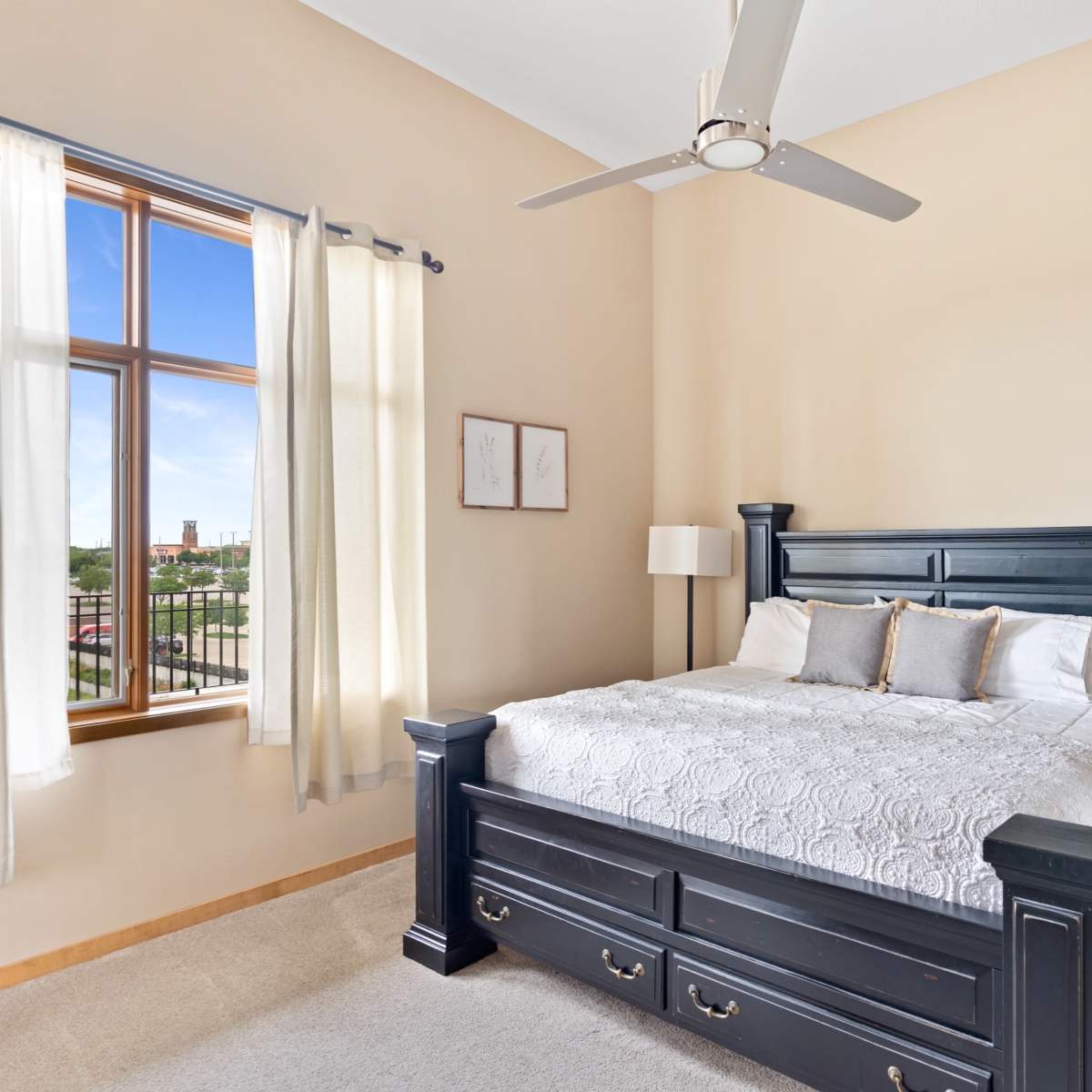 A well-lit bedroom with a dark wooden bed frame, a nightstand, and large windows allowing natural light to enter.