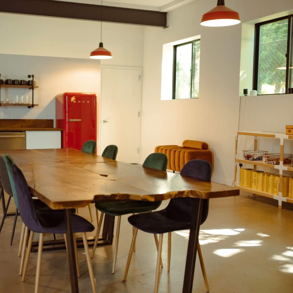 A spacious dining area features a long wooden table surrounded by colorful chairs, with a modern kitchen in the background and a red refrigerator.