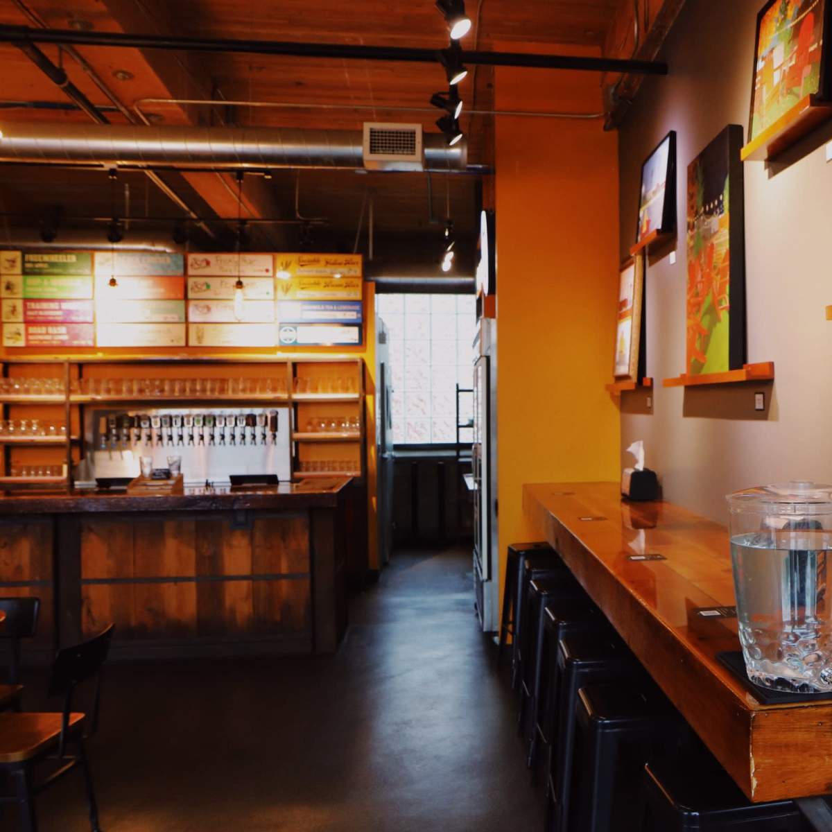The interior of a modern café featuring wooden shelves with colorful artwork, a long wooden counter, and a beverage station with glass pitchers and cups.