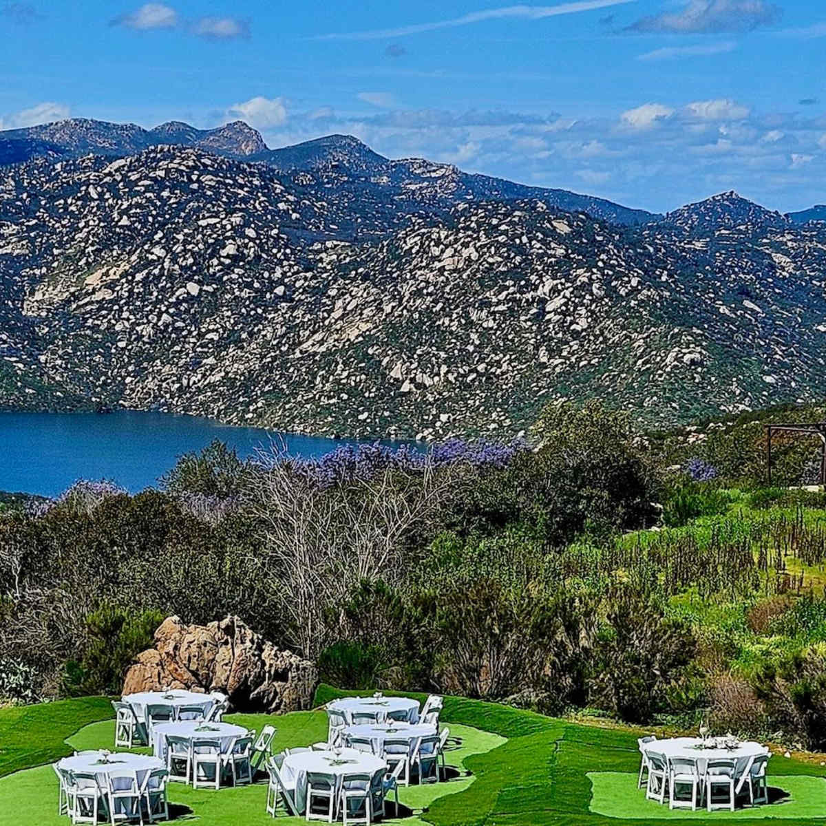 A scenic outdoor setting with white tables arranged on green grass, framed by mountains and a lake in the background.