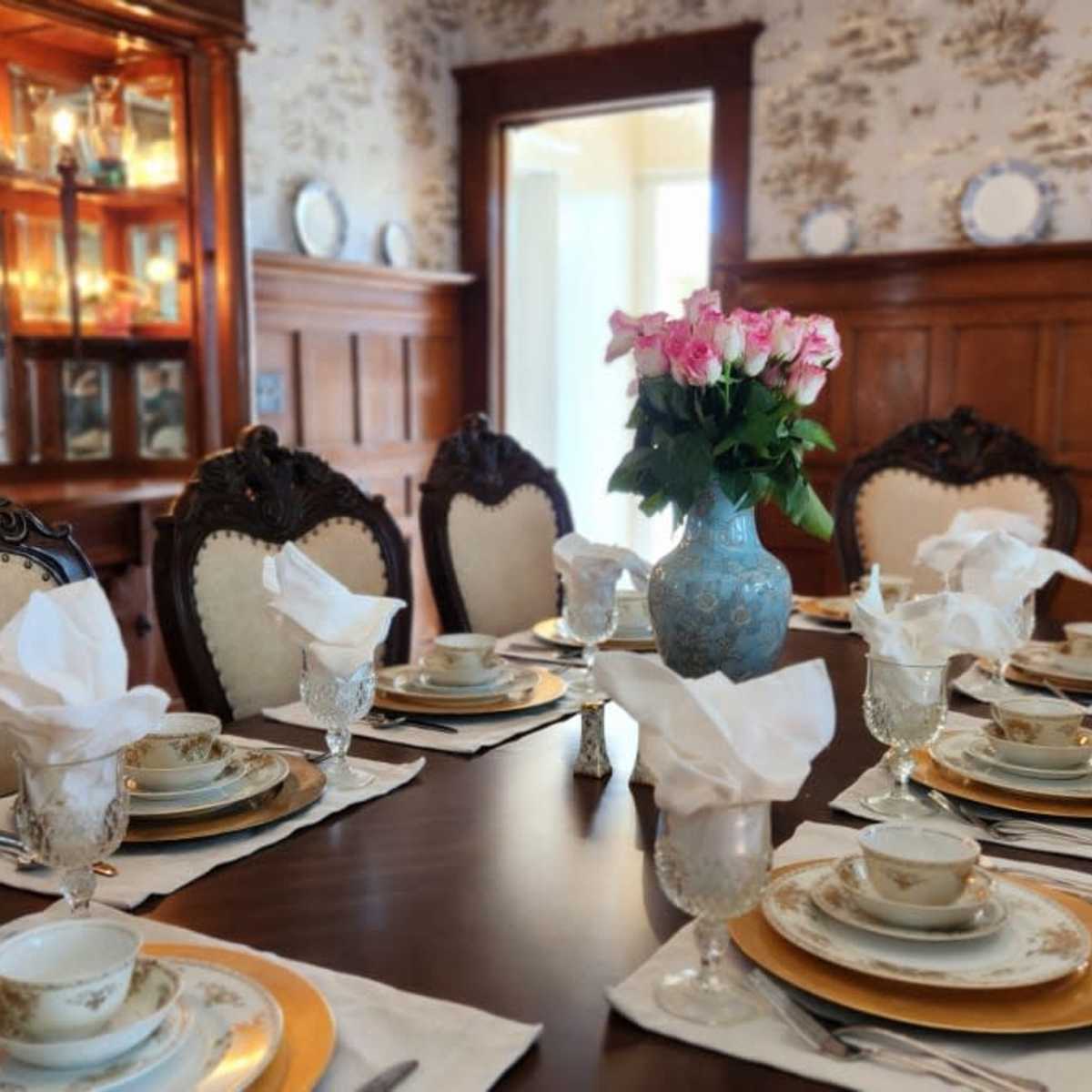 A dining room is set with an elegantly arranged table featuring fine china, glassware, and a vase of flowers, all surrounded by ornate wooden furnishings.