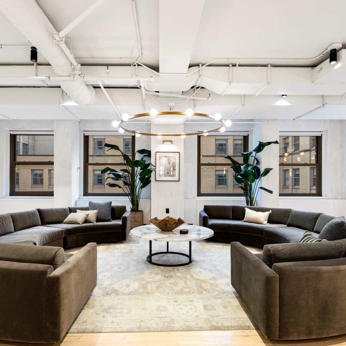A modern lounge area with two circular sofas facing a central coffee table, surrounded by large windows and indoor plants.