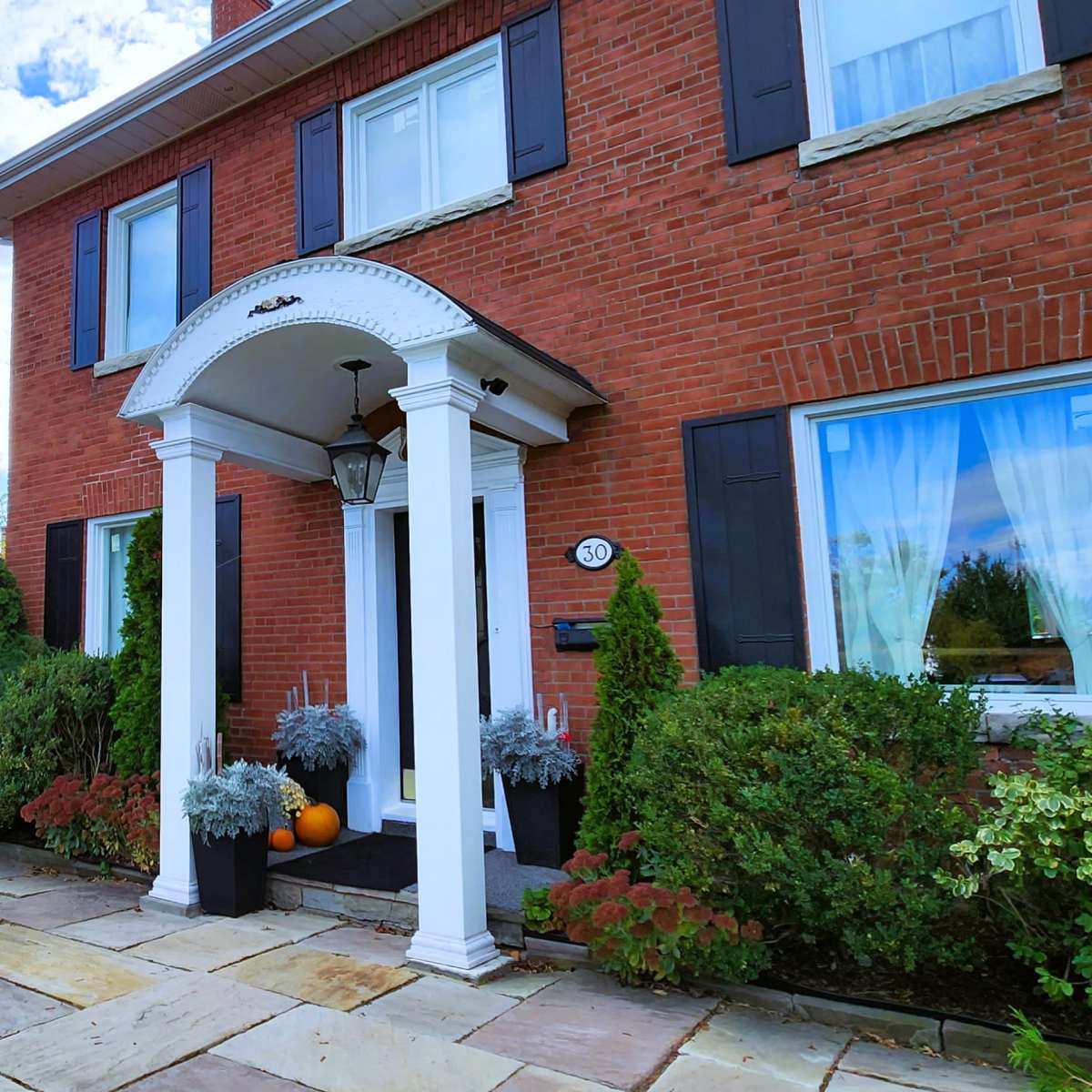 A brick house with white columns at the entrance, surrounded by greenery and a stone patio with two white chairs and potted plants.