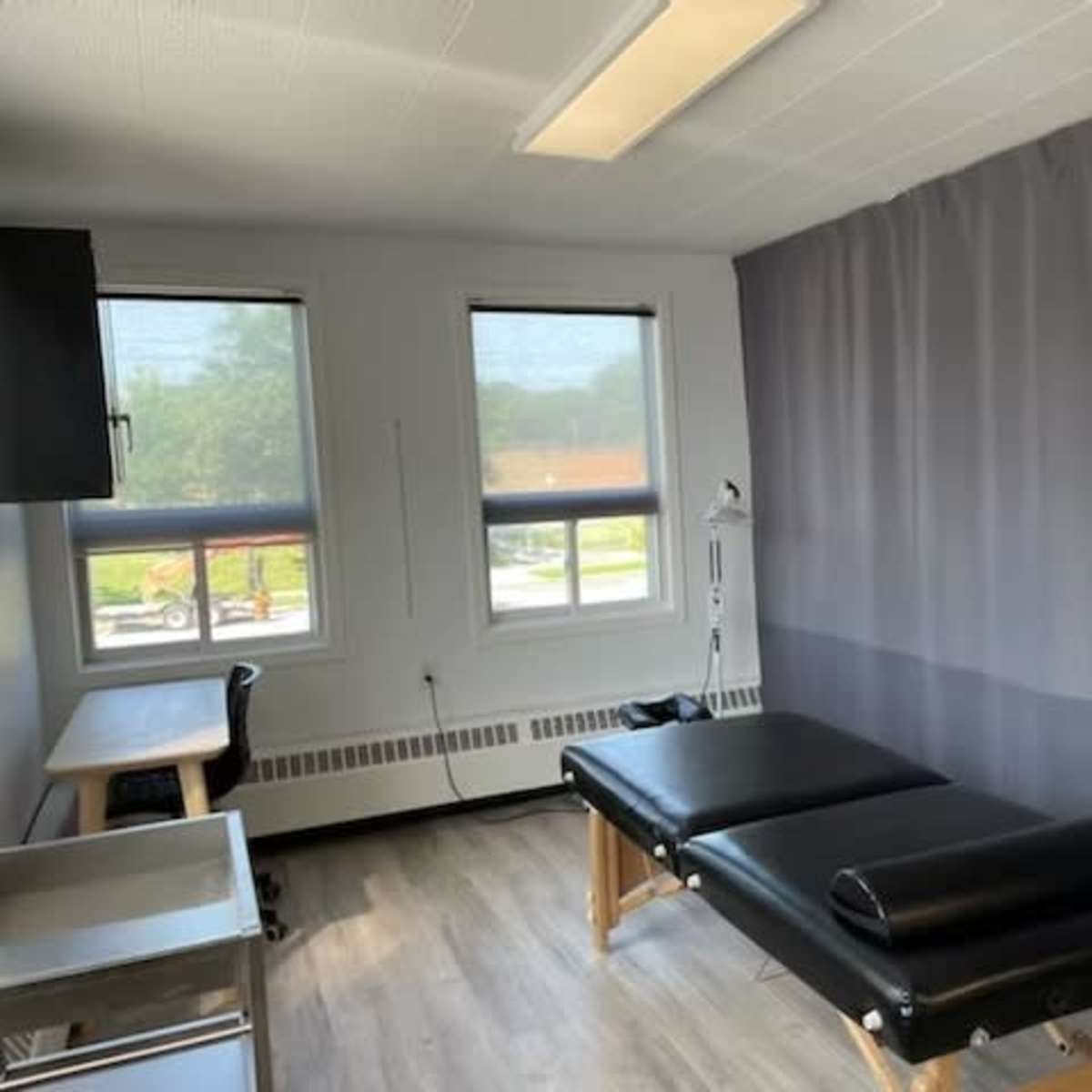 A treatment room with a black massage table, a desk, and large windows adorned with light curtains.