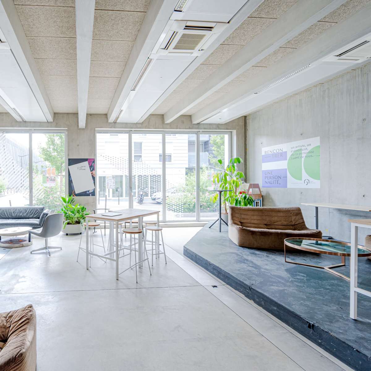 A modern workspace with concrete walls, large windows letting in natural light, and a mix of seating arrangements including chairs, tables, and plants.