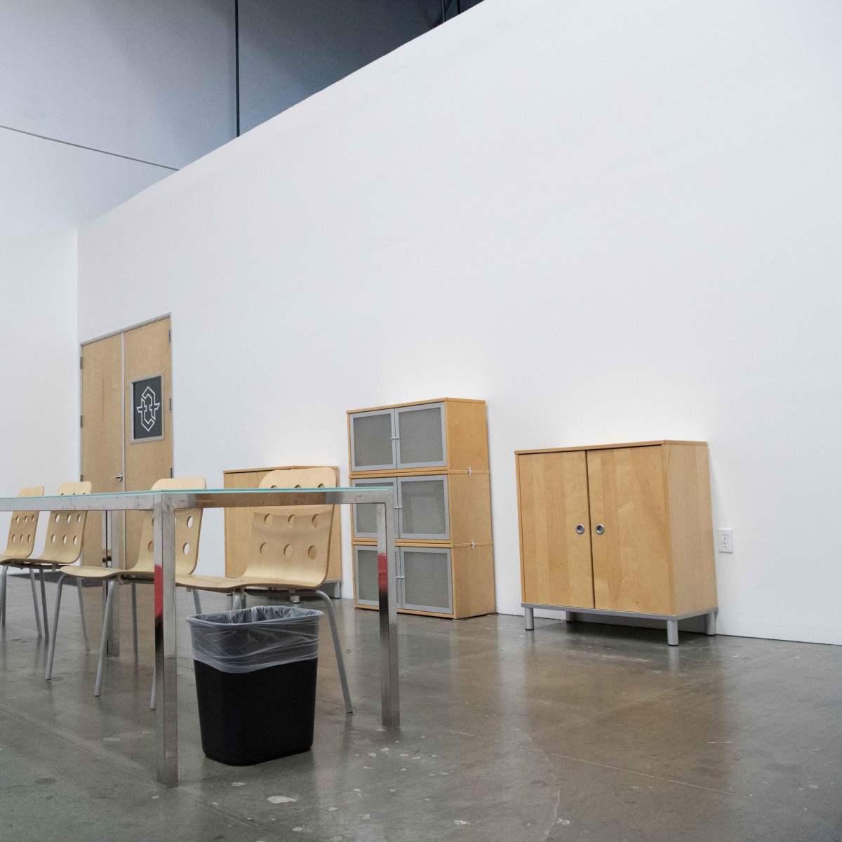 An empty room features a long table with four chairs, a trash bin underneath, and wooden cabinets against the wall.