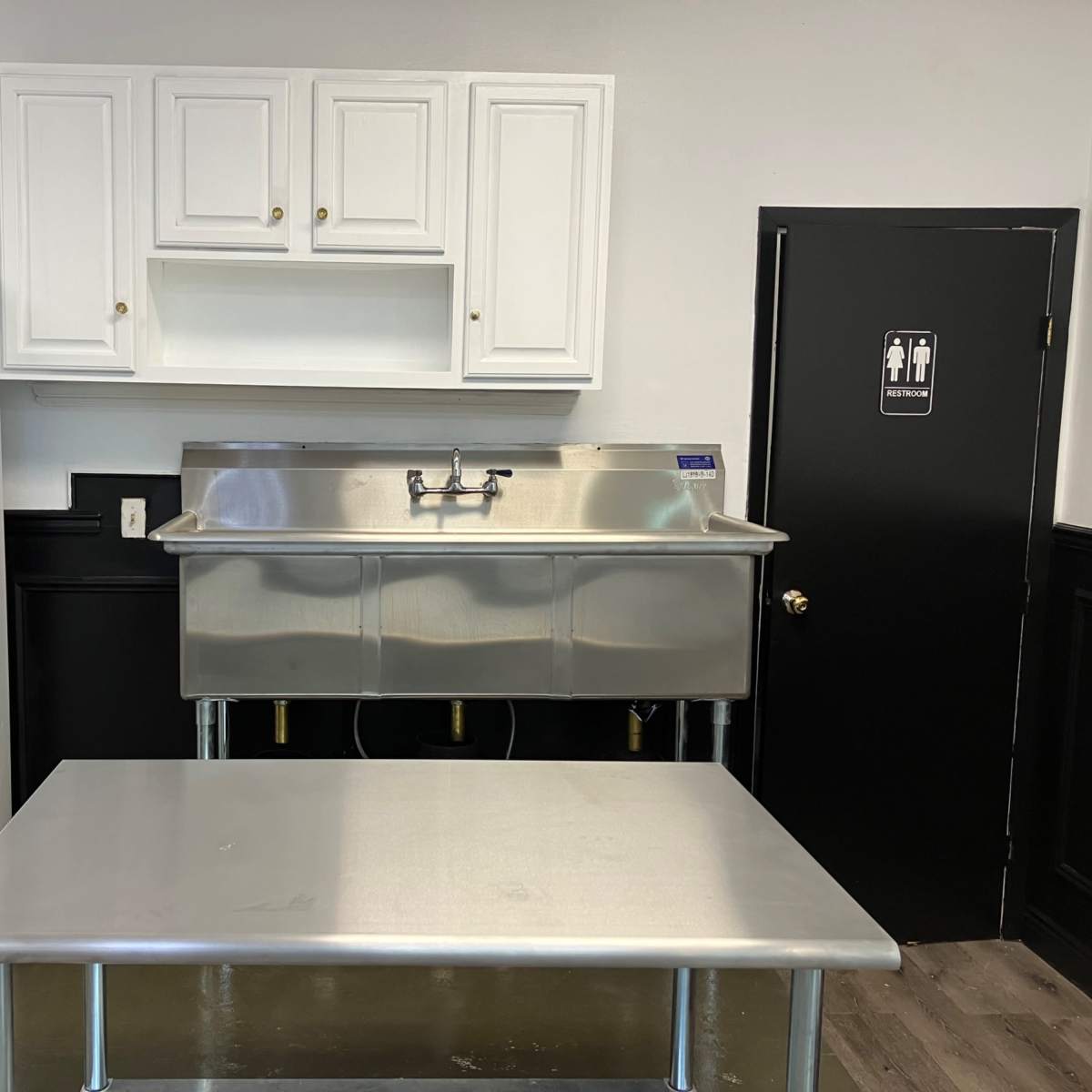 A commercial kitchen space featuring a stainless steel sink with a countertop, a white refrigerator, and black-and-white decor, including a restroom door.