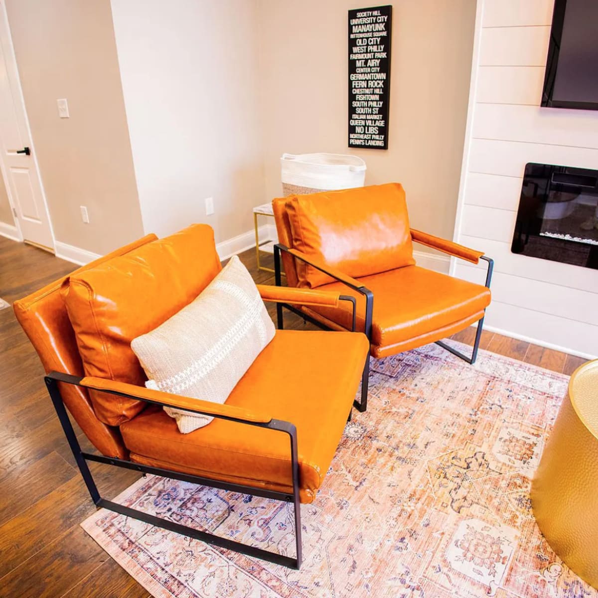 Two orange chairs are positioned on a rug in a modern living room, accompanied by a gold coffee table and a wall-mounted television.