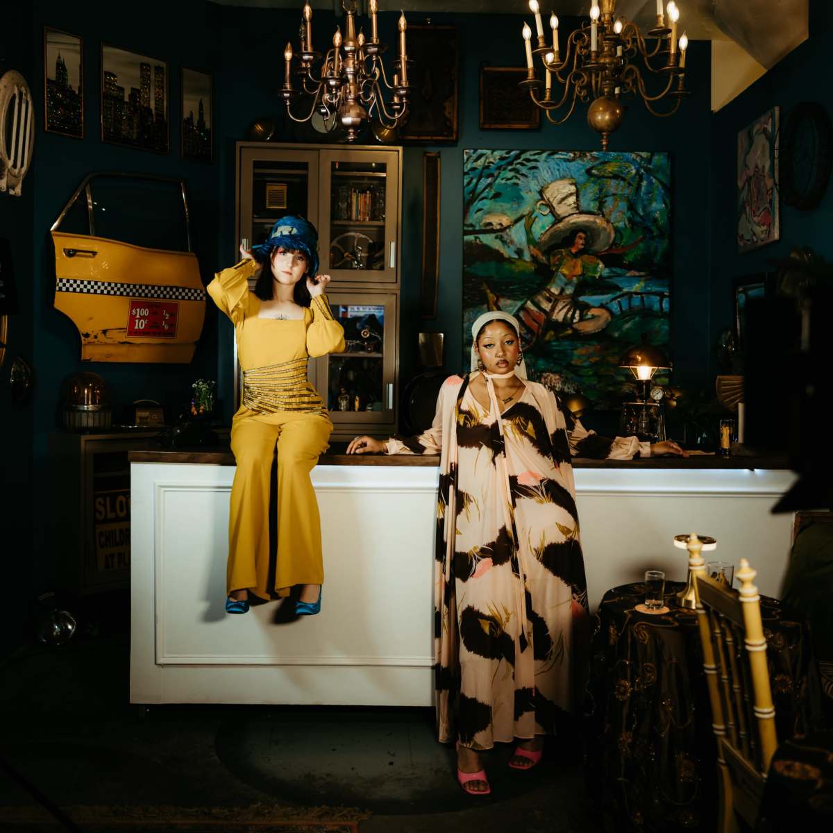Two women dressed in vibrant outfits pose inside a stylish interior featuring art, chandeliers, and eclectic decor.