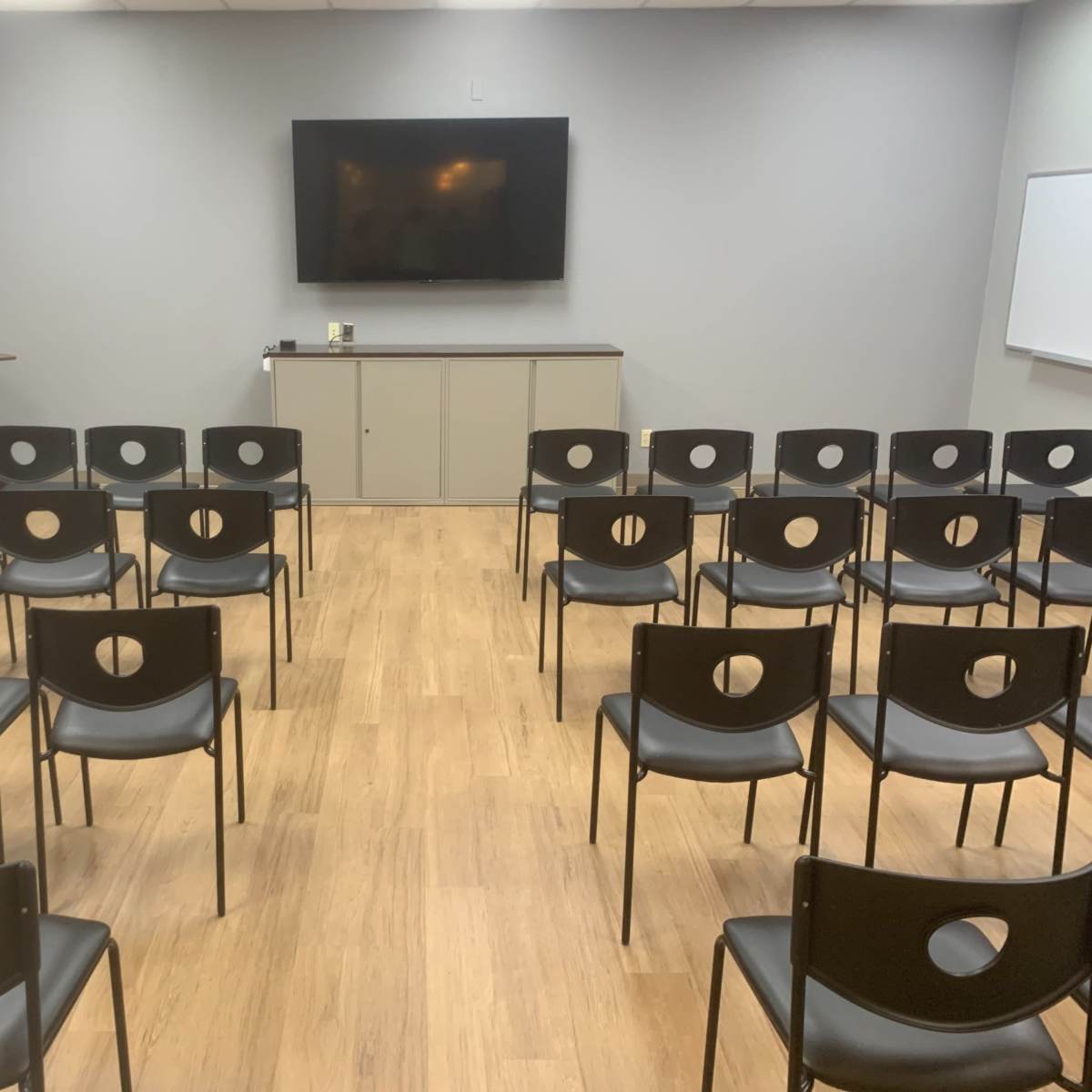 A meeting room is set up with rows of black chairs facing a wall-mounted television and a whiteboard.