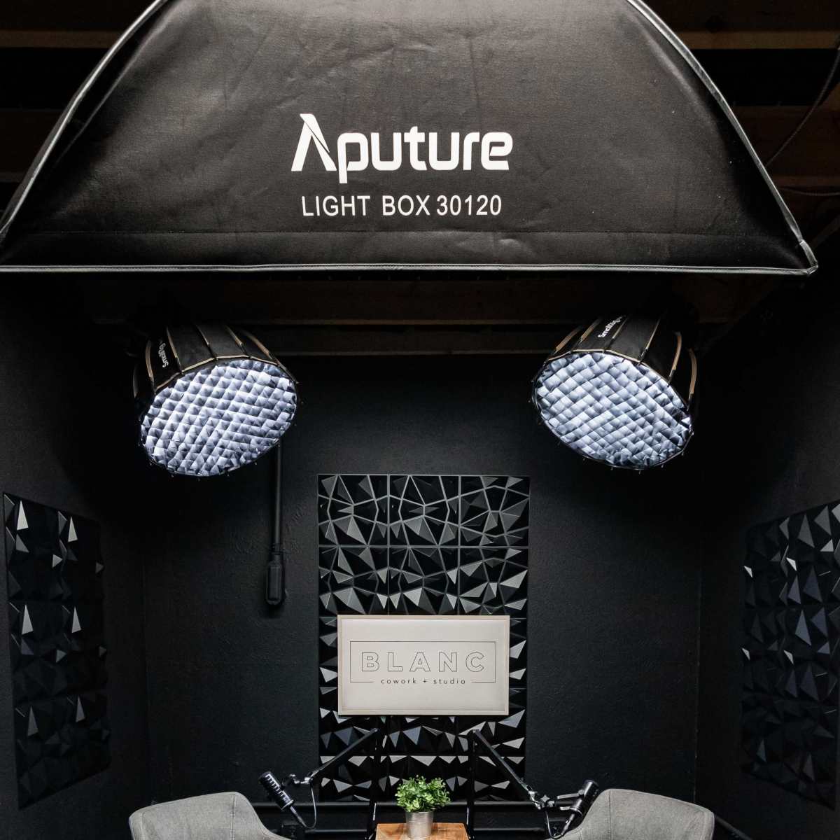 A photography setup features two gray chairs and a wooden table under a large lightbox, with geometric black wall panels in a dimly lit environment.