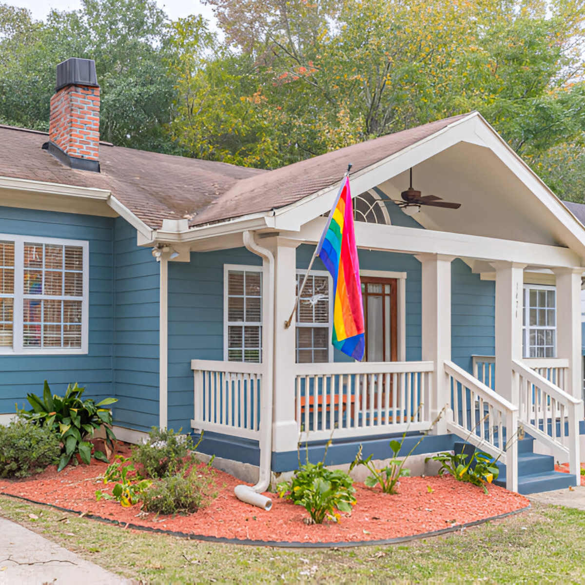 A blue house with a front porch, a rainbow flag hanging from the porch, and landscaped flower beds in the yard.