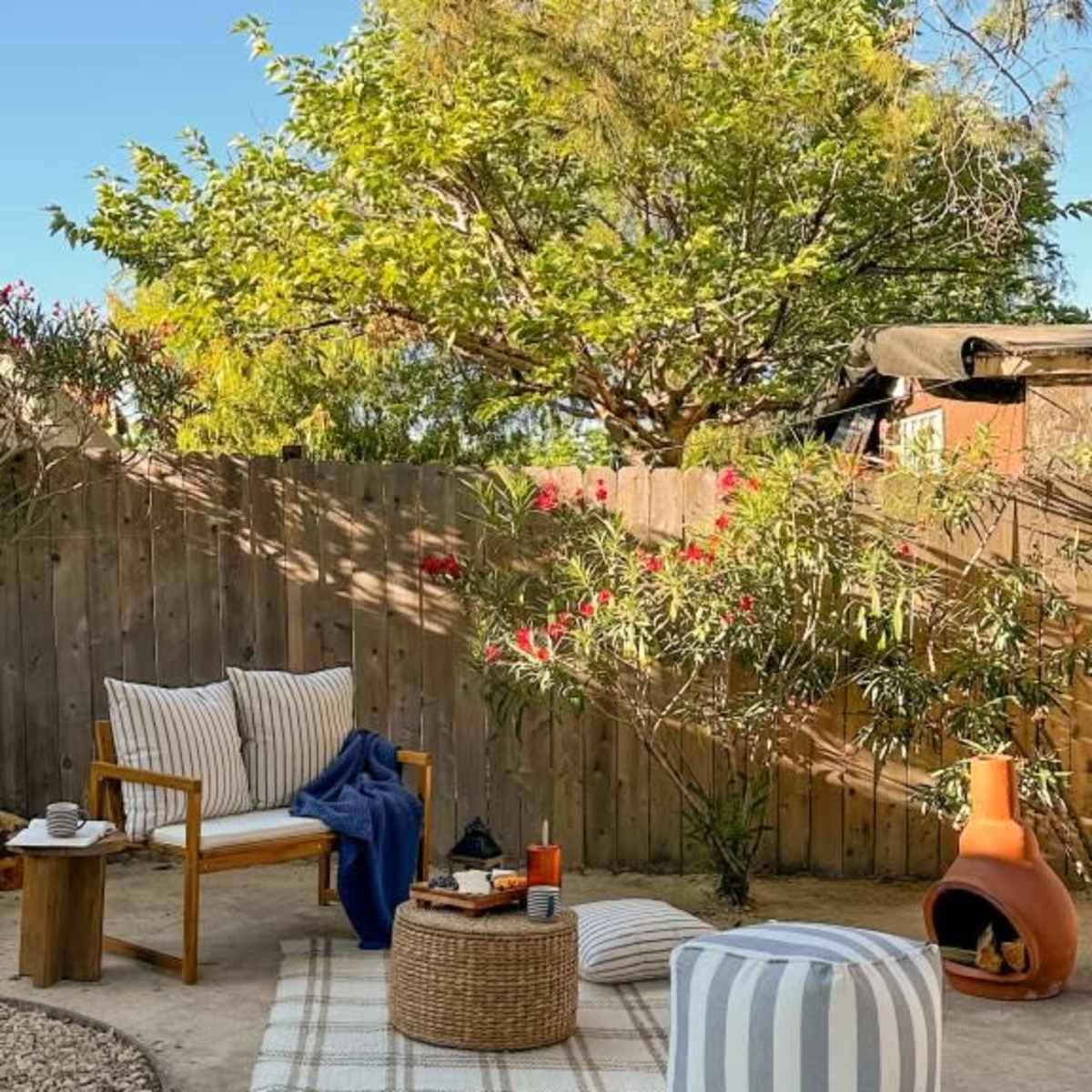 A cozy outdoor seating area features a wooden chair, a round woven table, and a striped pouf, surrounded by plants and a fence.