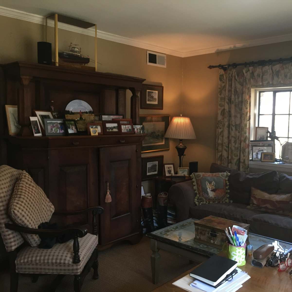 A cozy living room featuring a dark wooden cabinet, a comfortable sofa with decorative cushions, and a window adorned with patterned curtains, all arranged around a central glass coffee table.
