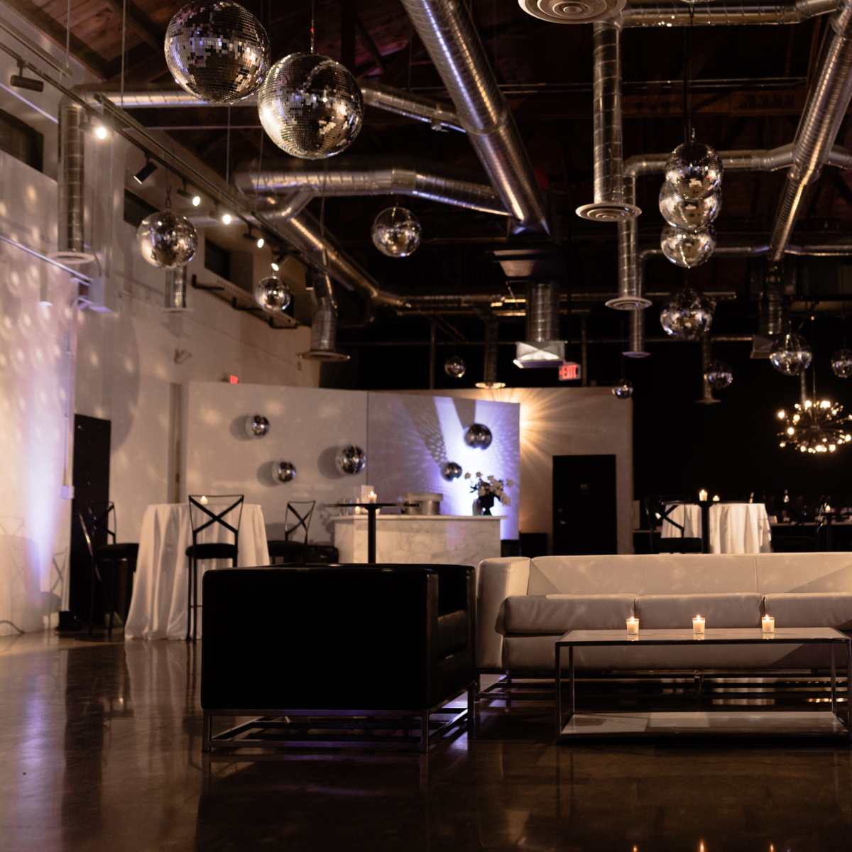 A modern event space featuring lounge seating, disco balls hanging from the ceiling, and a bar area with tables in a stylishly lit environment.