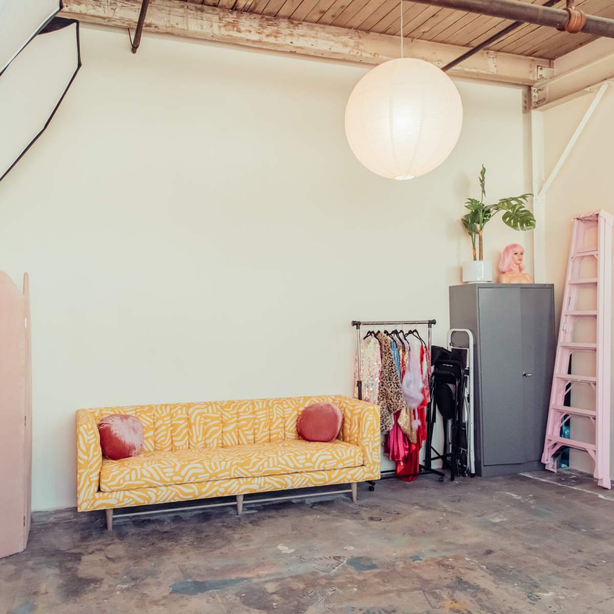 A minimalist studio space featuring a yellow patterned sofa, a clothing rack, a gray cabinet, a pink ladder, and a round paper lantern hanging from the ceiling.