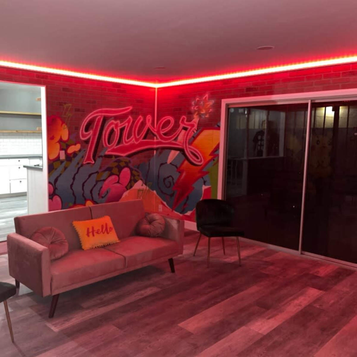 A modern living space with a pink sofa, two chairs, and a colorful mural on a brick wall, illuminated by red LED lights.