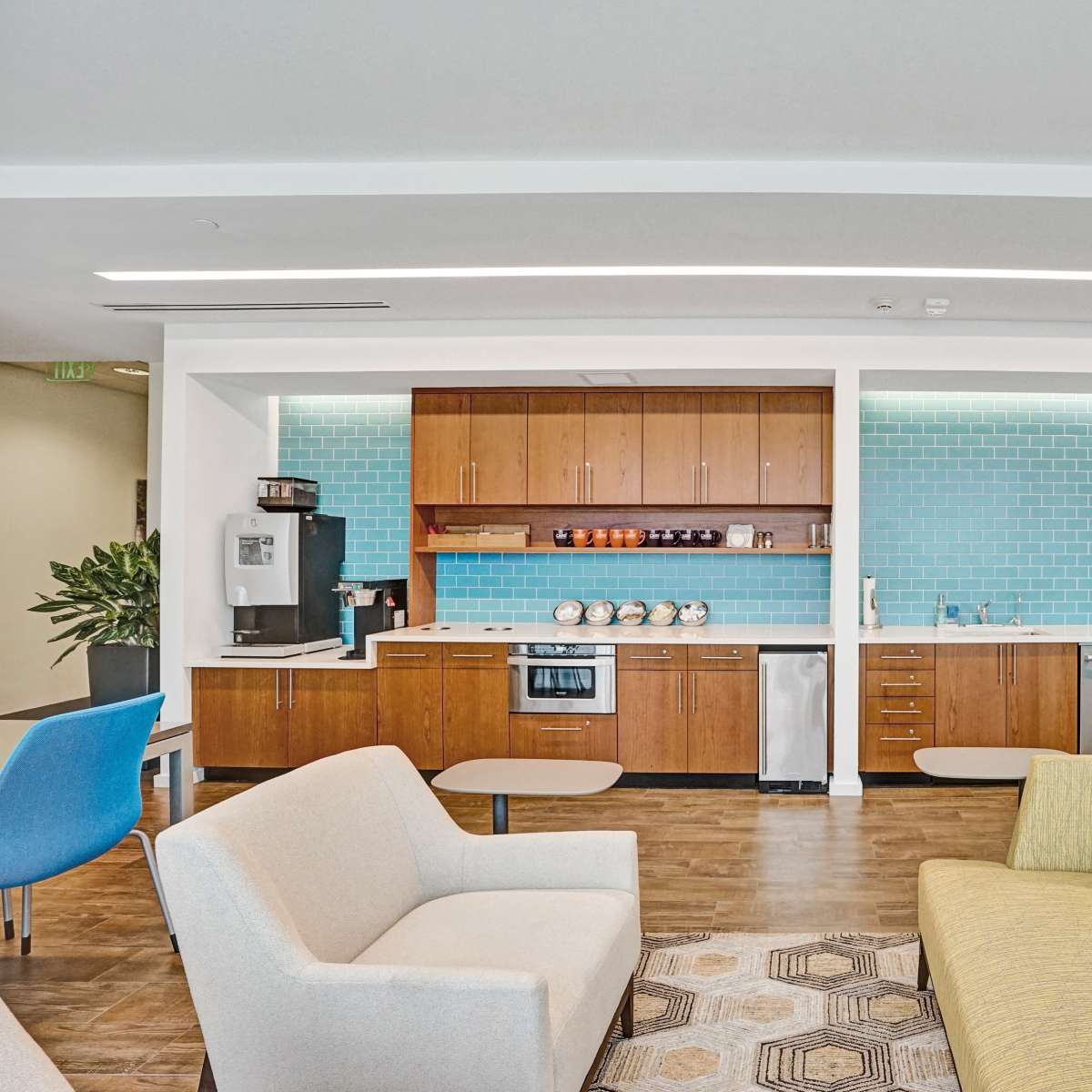 A modern kitchen area featuring wooden cabinets, a stainless steel refrigerator, and a blue tile backsplash, accompanied by a lounge space with a sofa and chairs.