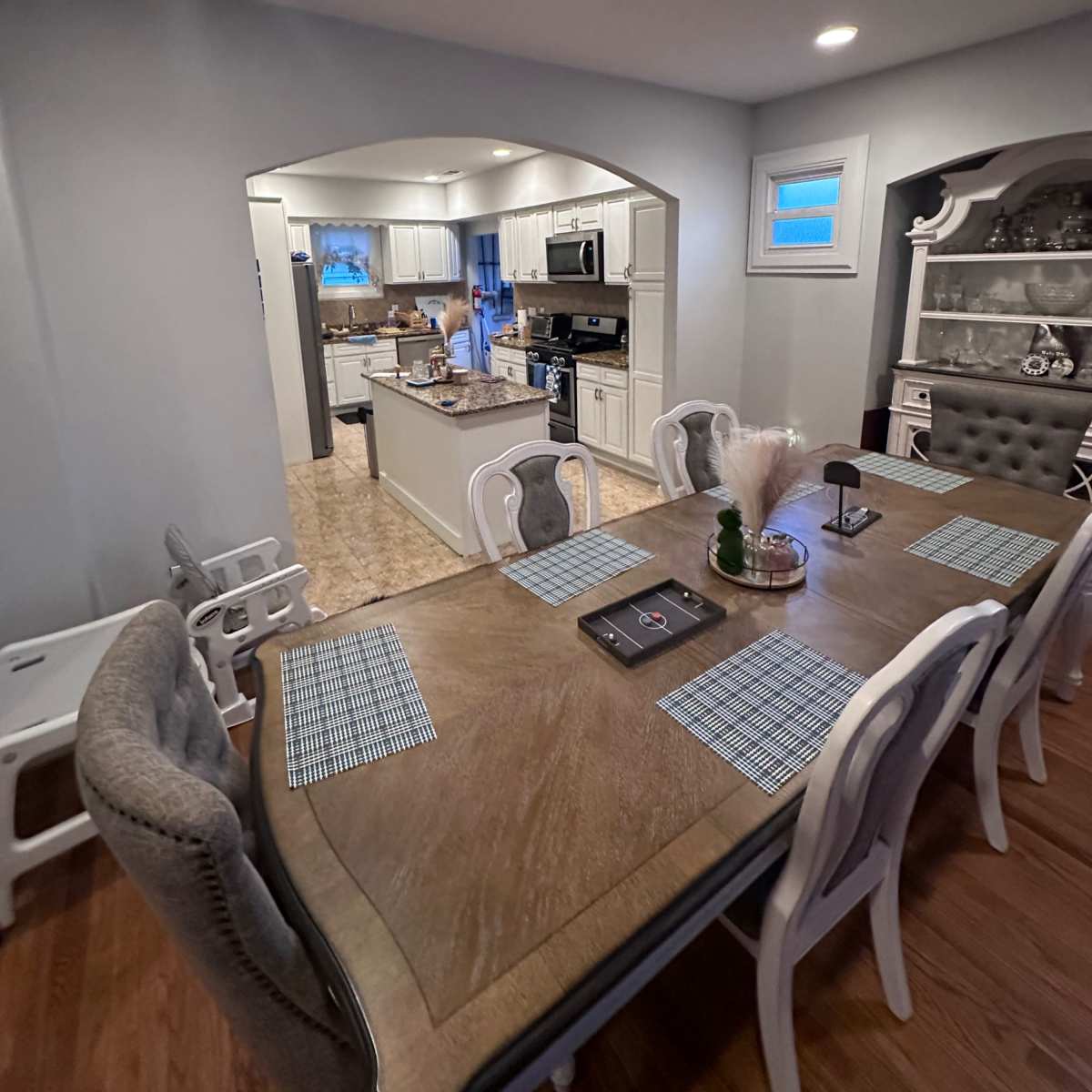A dining area with a wooden table set for a meal, leading into a kitchen visible in the background.