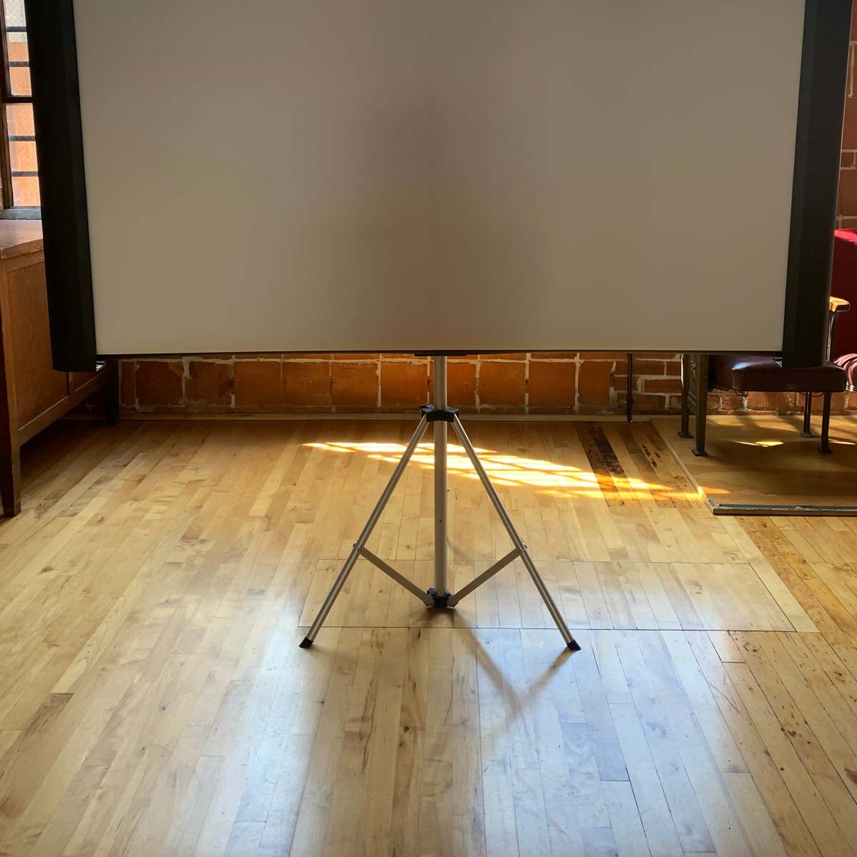 A large, blank projection screen set up on a tripod in a room with wooden floors and brick walls.