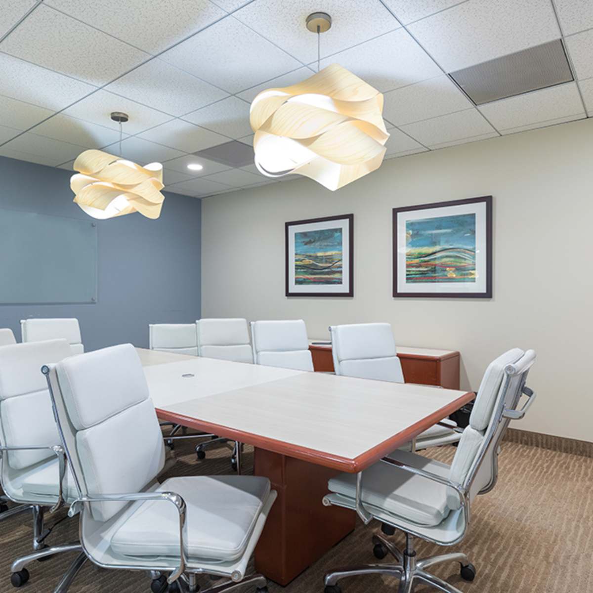 A modern conference room with a long table surrounded by white leather chairs, and two abstract art pieces hanging on the wall.