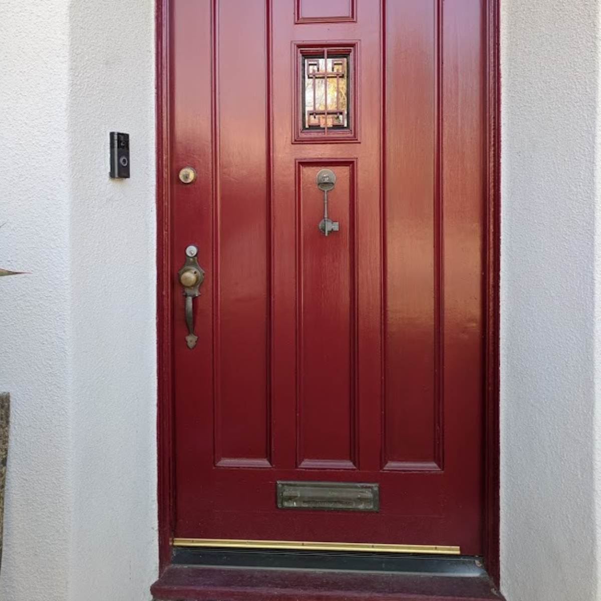 A red wooden door with a decorative window and a brass doorknob, set into a white wall.