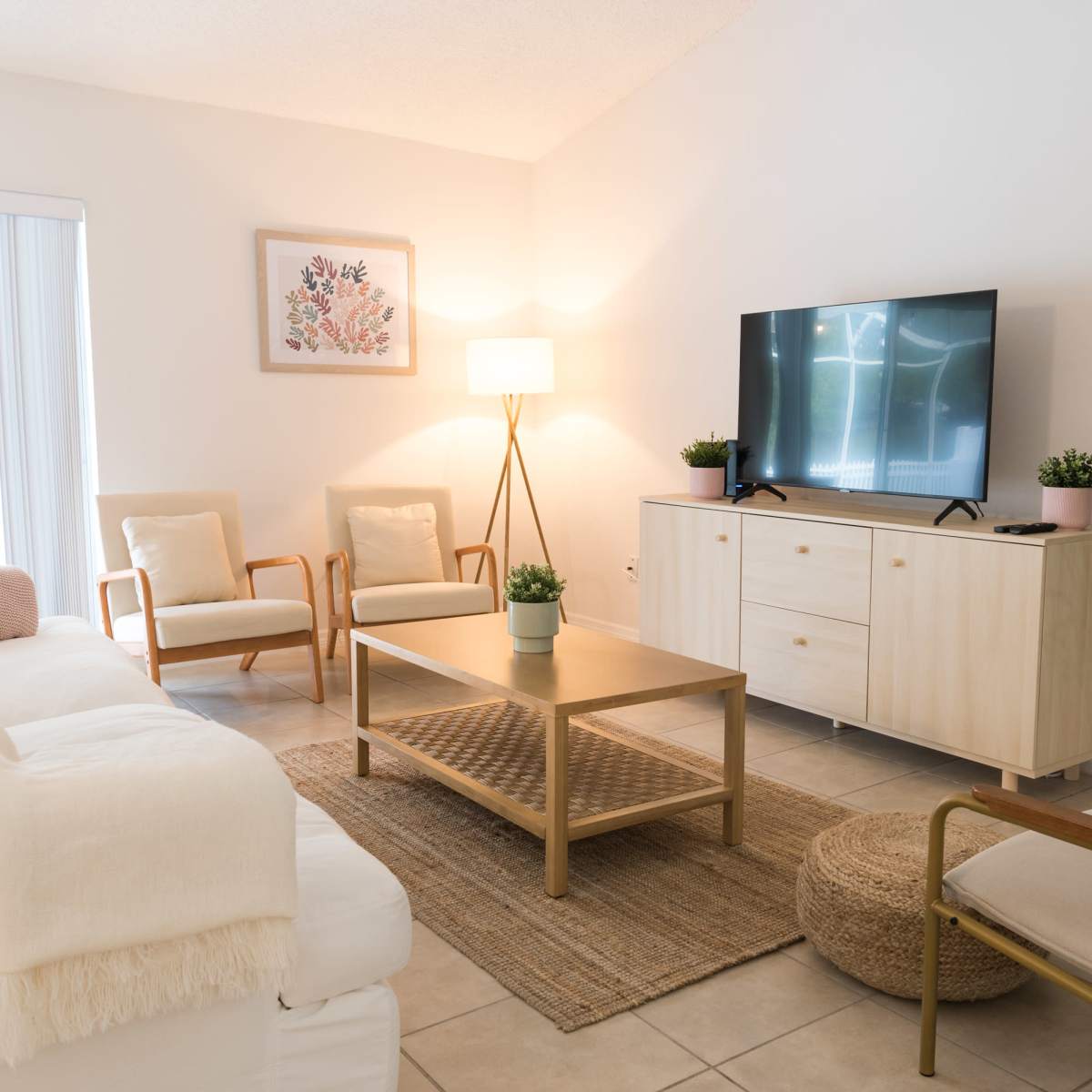 A modern living room features a light-colored sofa, two armchairs, a coffee table, a media console with a television, and decorative plants.