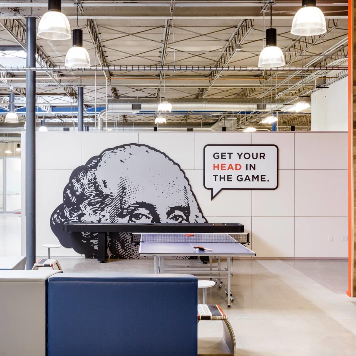 A modern office space featuring a ping pong table, lounge seating, and a wall graphic of George Washington with the phrase "GET YOUR HEAD IN THE GAME."