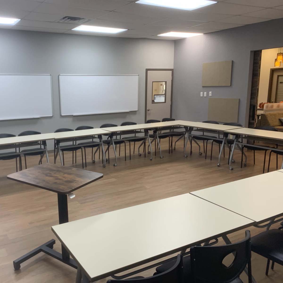 A classroom with a circular arrangement of tables and chairs, two whiteboards on the front wall, and a flat-screen monitor.