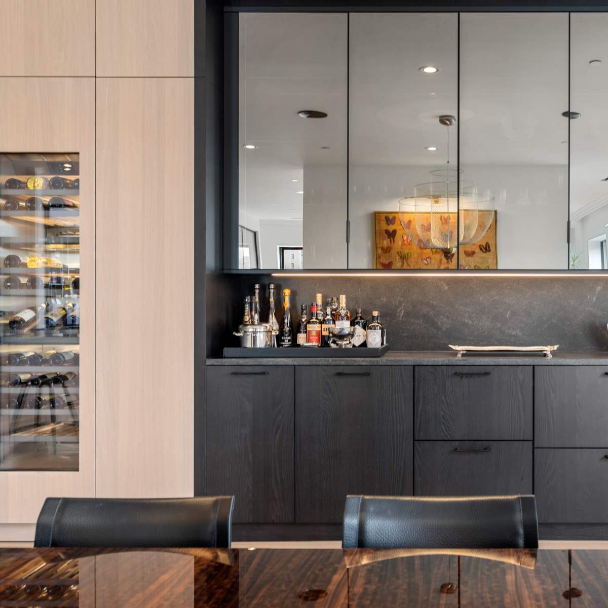 A modern kitchen area featuring a black cabinetry wall with a mirrored backsplash, a countertop displaying various bottles, and a wine cooler to the left.