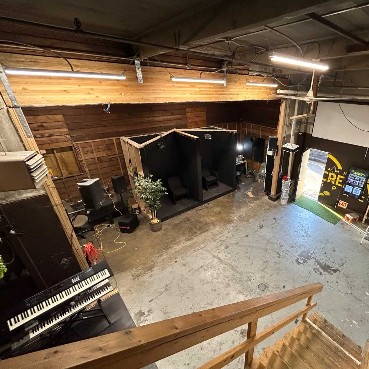 A spacious interior view of a creative workspace featuring a piano, black cubicles, and a projector screen against wooden walls.