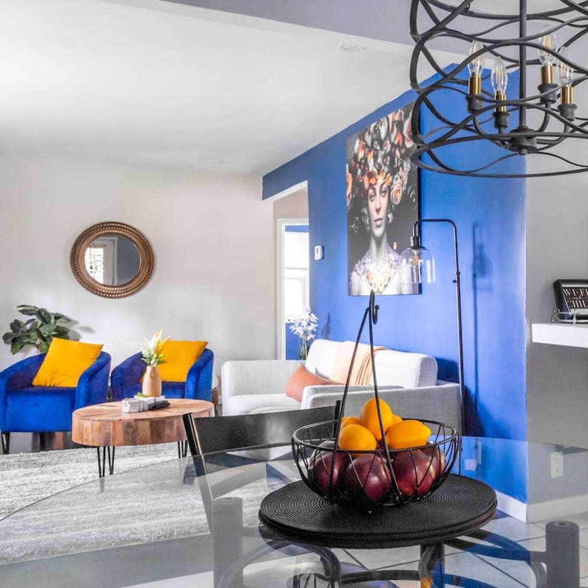A modern living space features a round dining table with a fruit bowl, a gray sofa, blue accent chairs, and vibrant wall art.