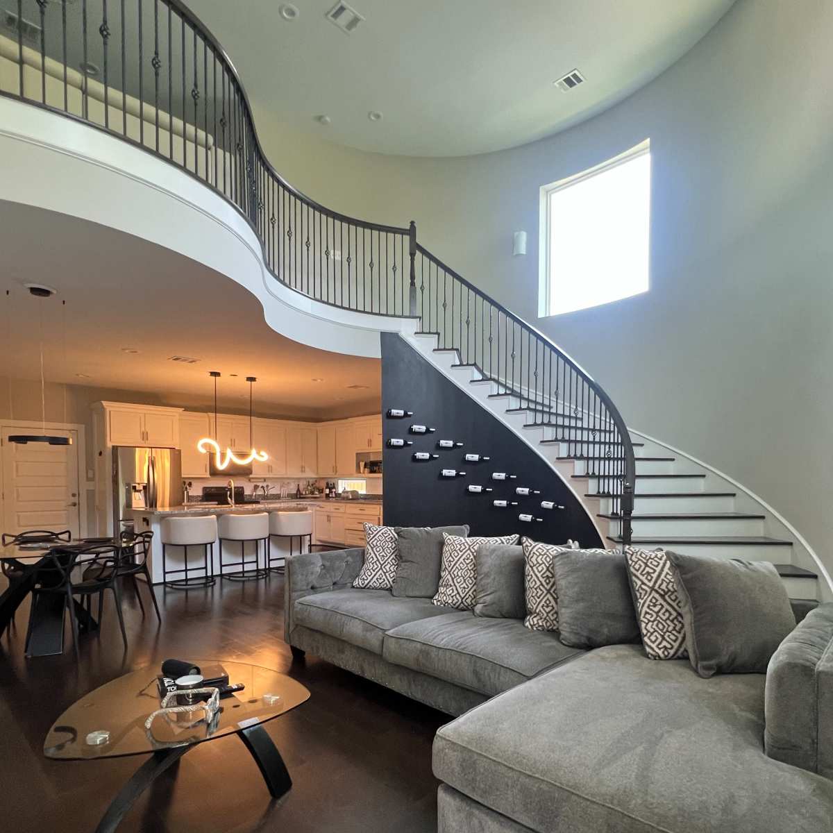 A modern living room with a large gray sectional sofa, a coffee table, and a sweeping staircase that leads to an upper floor.