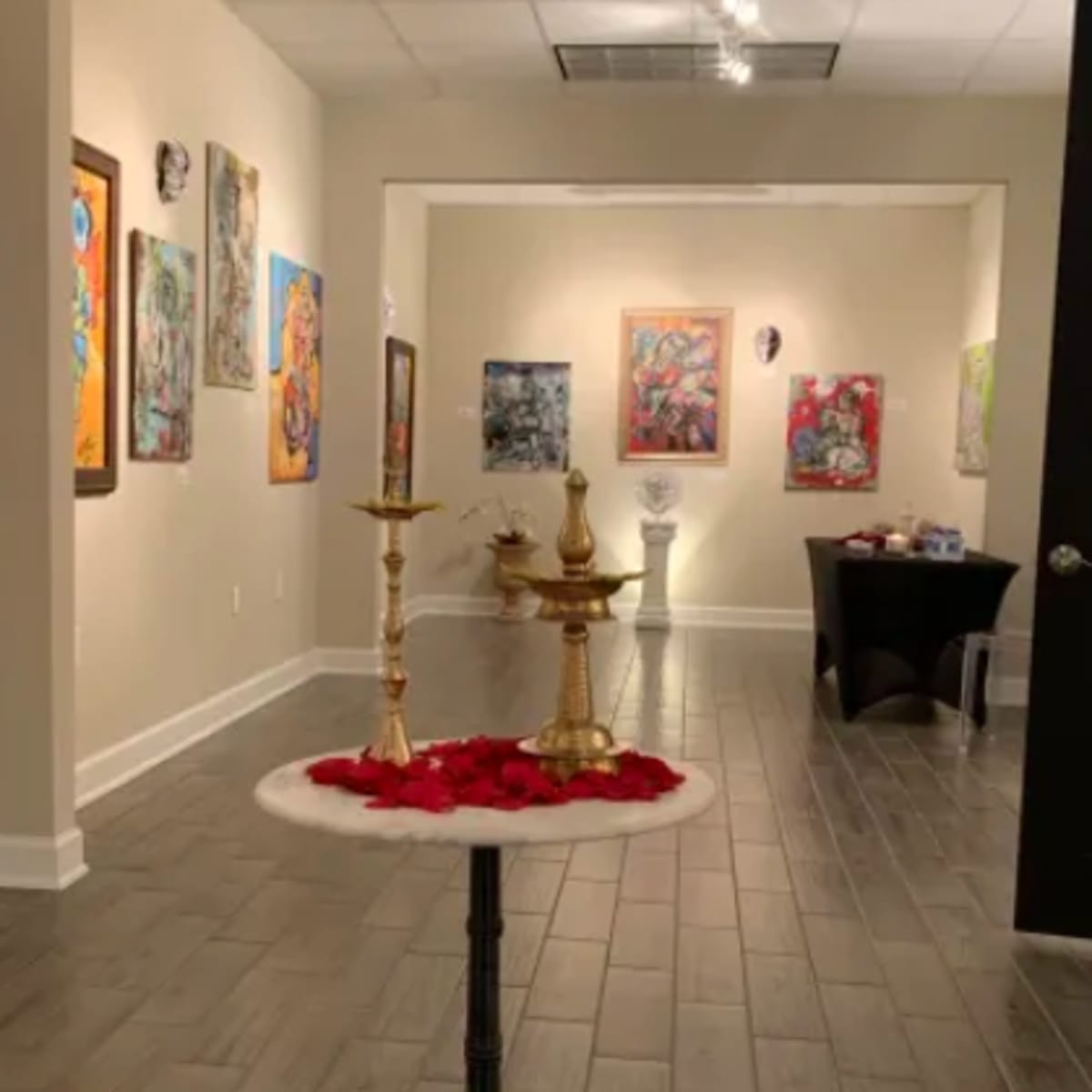 An art gallery adorned with colorful paintings on the walls, featuring a small table with decorative items and rose petals in the foreground.