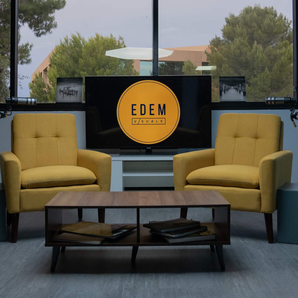 A modern studio setup with two yellow chairs facing a coffee table, a television displaying the logo "EDEM," and large windows with greenery outside.