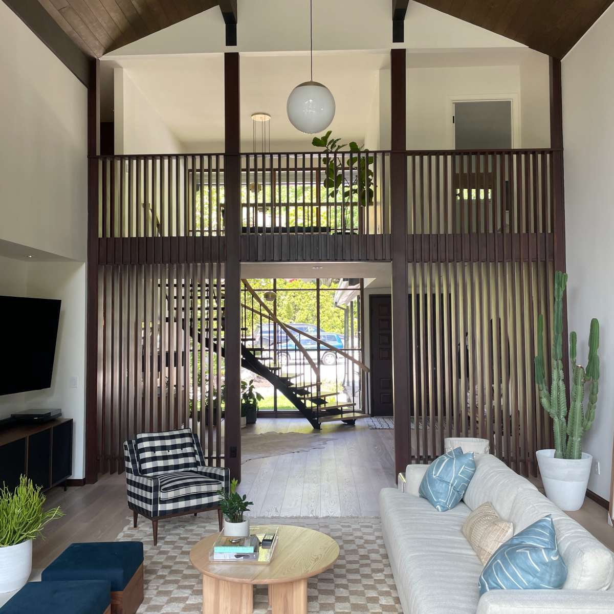 A modern living room with a high ceiling, featuring a sofa, a circular coffee table, a patterned rug, and a staircase visible in the background, separated by slatted wooden partitions.