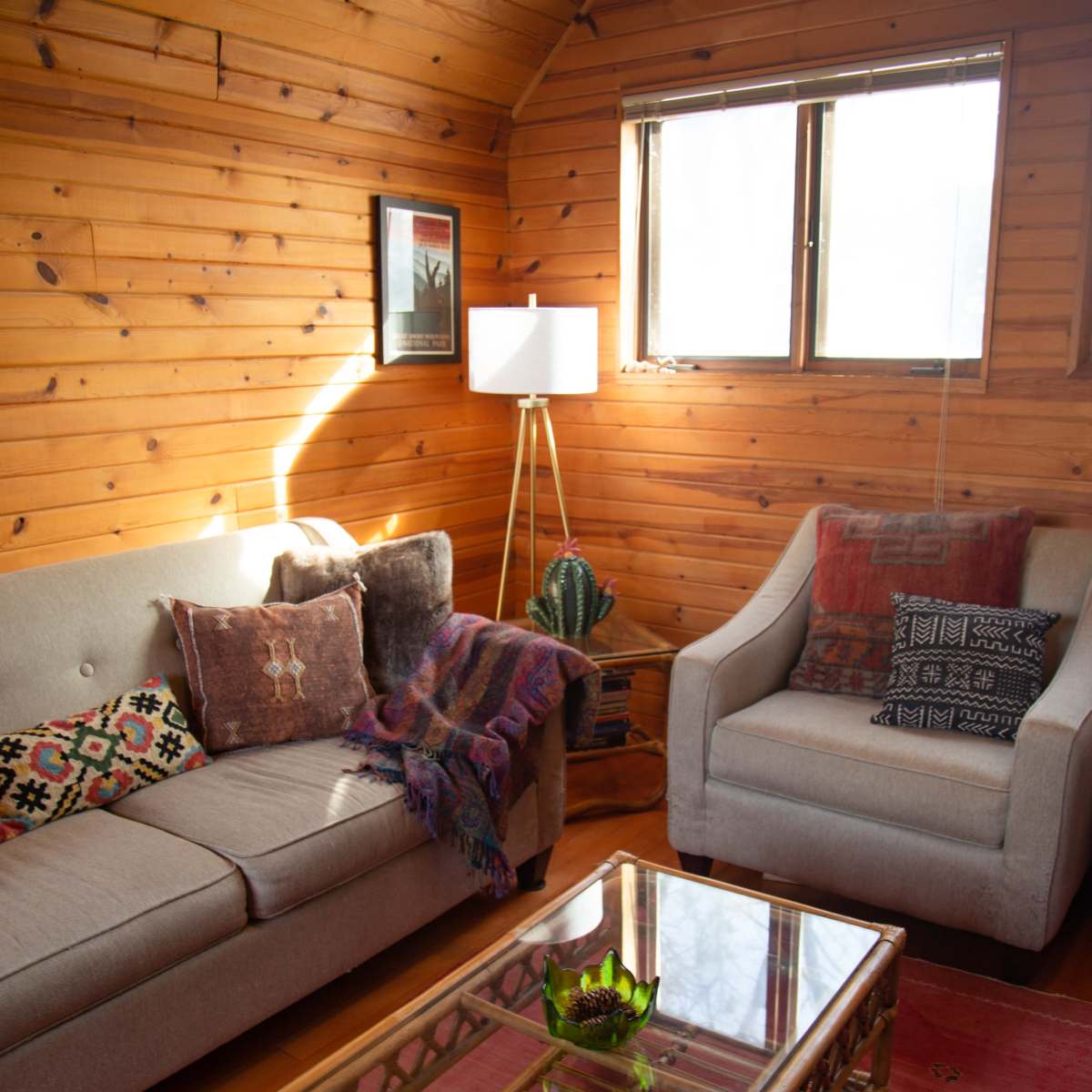 The cozy living room features a beige sofa and a light-colored armchair, surrounded by wooden walls, a glass coffee table, and a floor lamp.