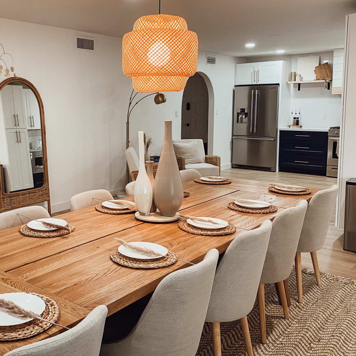 A dining room features a long wooden table set with plates and woven placemats, illuminated by a large woven pendant light, with a mirror and a modern kitchen visible in the background.