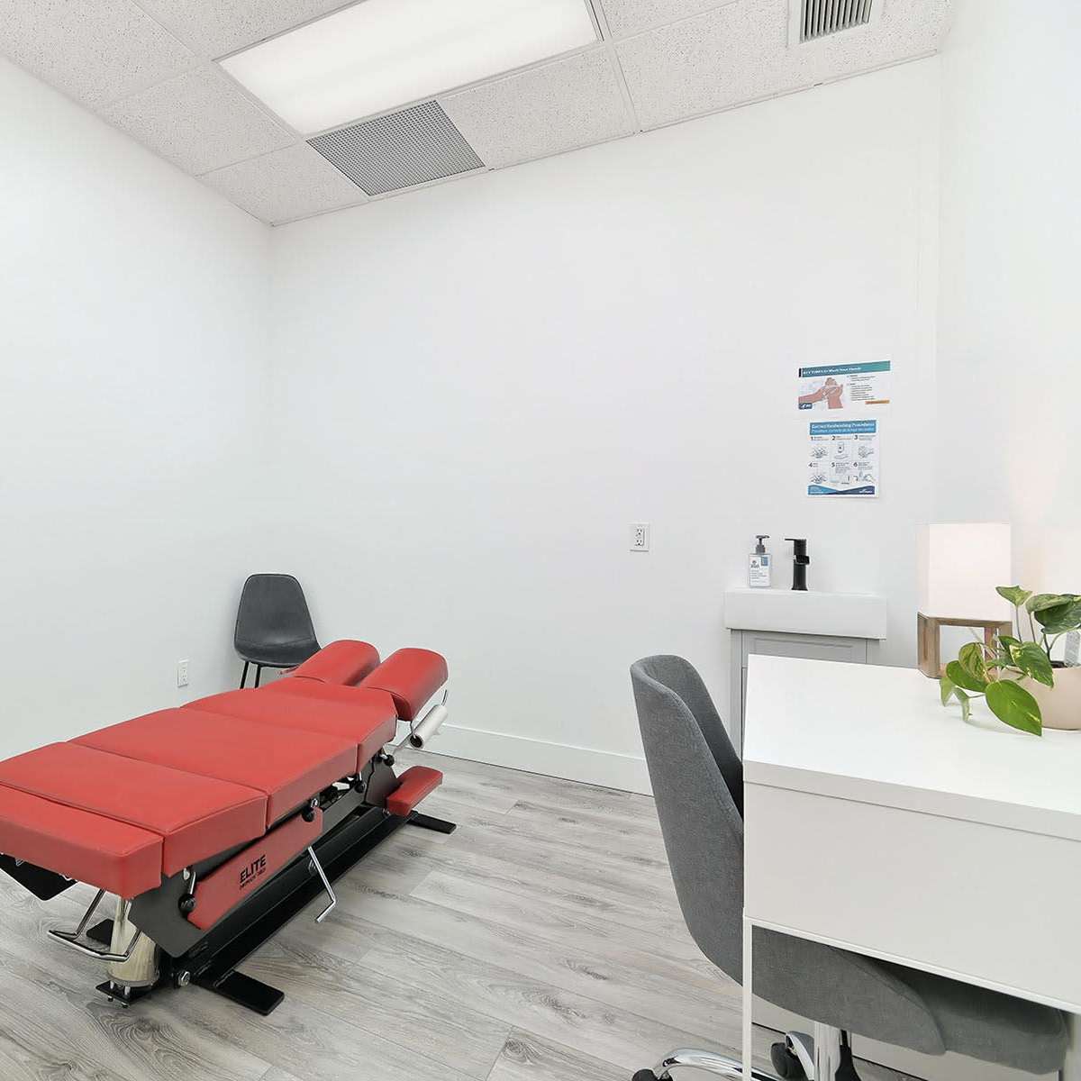 A clean therapy room featuring a red treatment table, a desk with a plant, and a gray chair.
