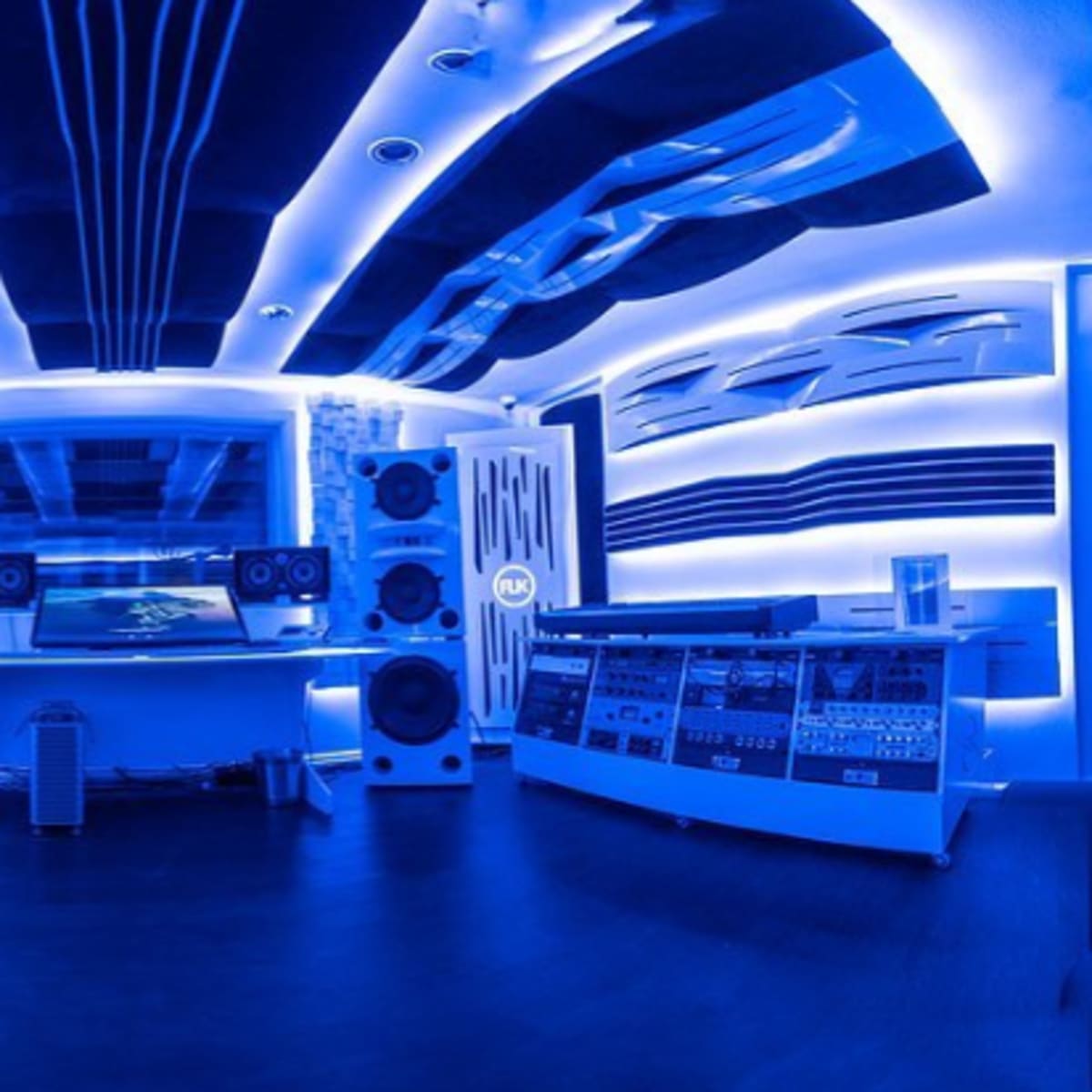 A modern music studio with blue ambient lighting, featuring large speakers and soundproofing panels on the walls.