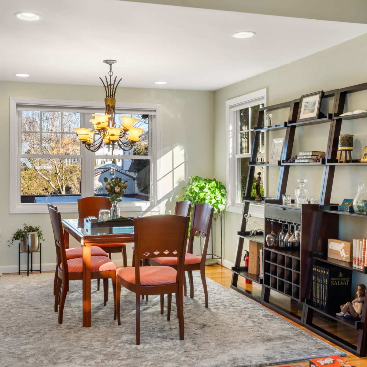 A well-lit dining area featuring a wooden table surrounded by four chairs, a bookshelf filled with various items, and large windows allowing natural light to enter.
