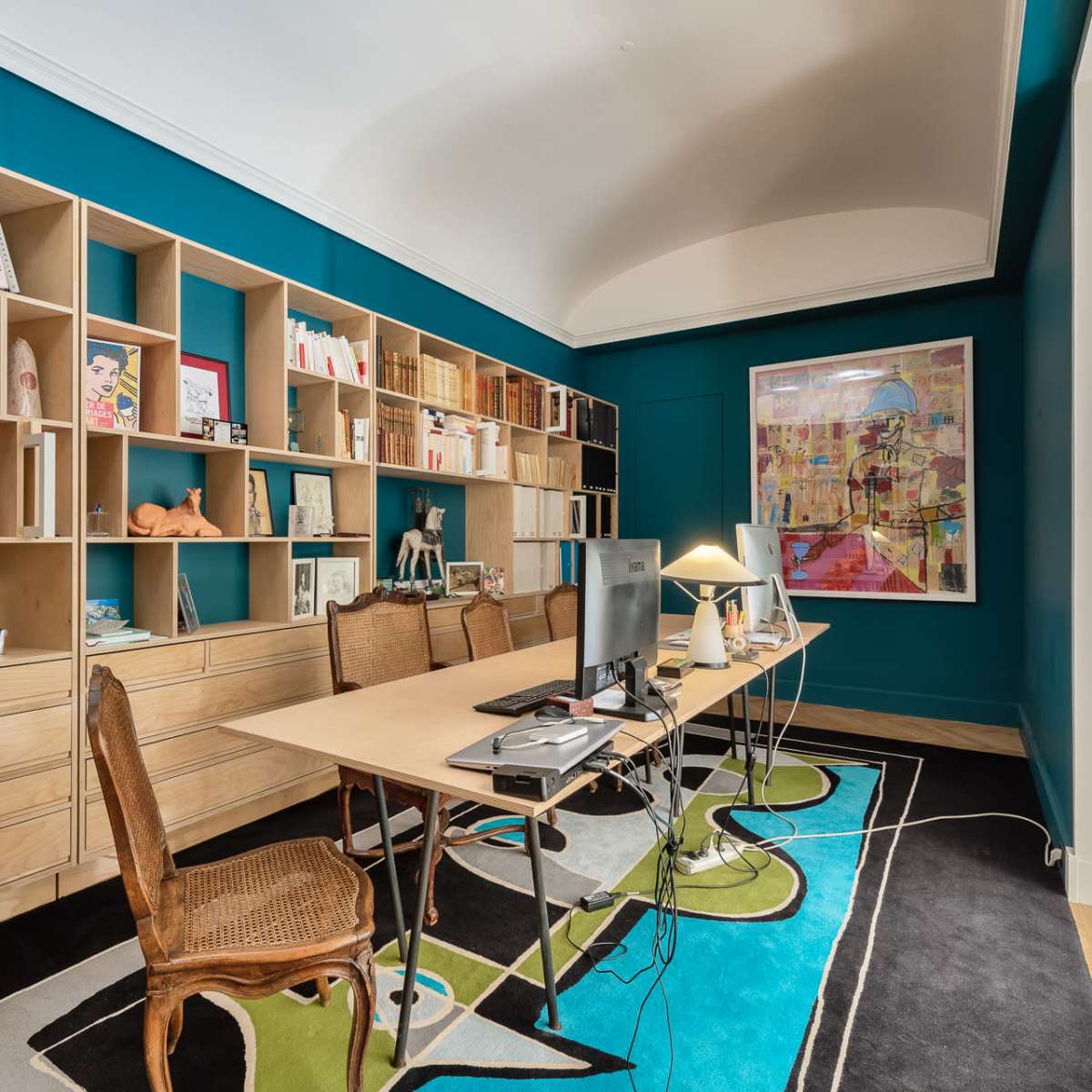 A home office with a long table in the center, flanked by two chairs, and a large bookshelf filled with books and decorative items against a teal wall.