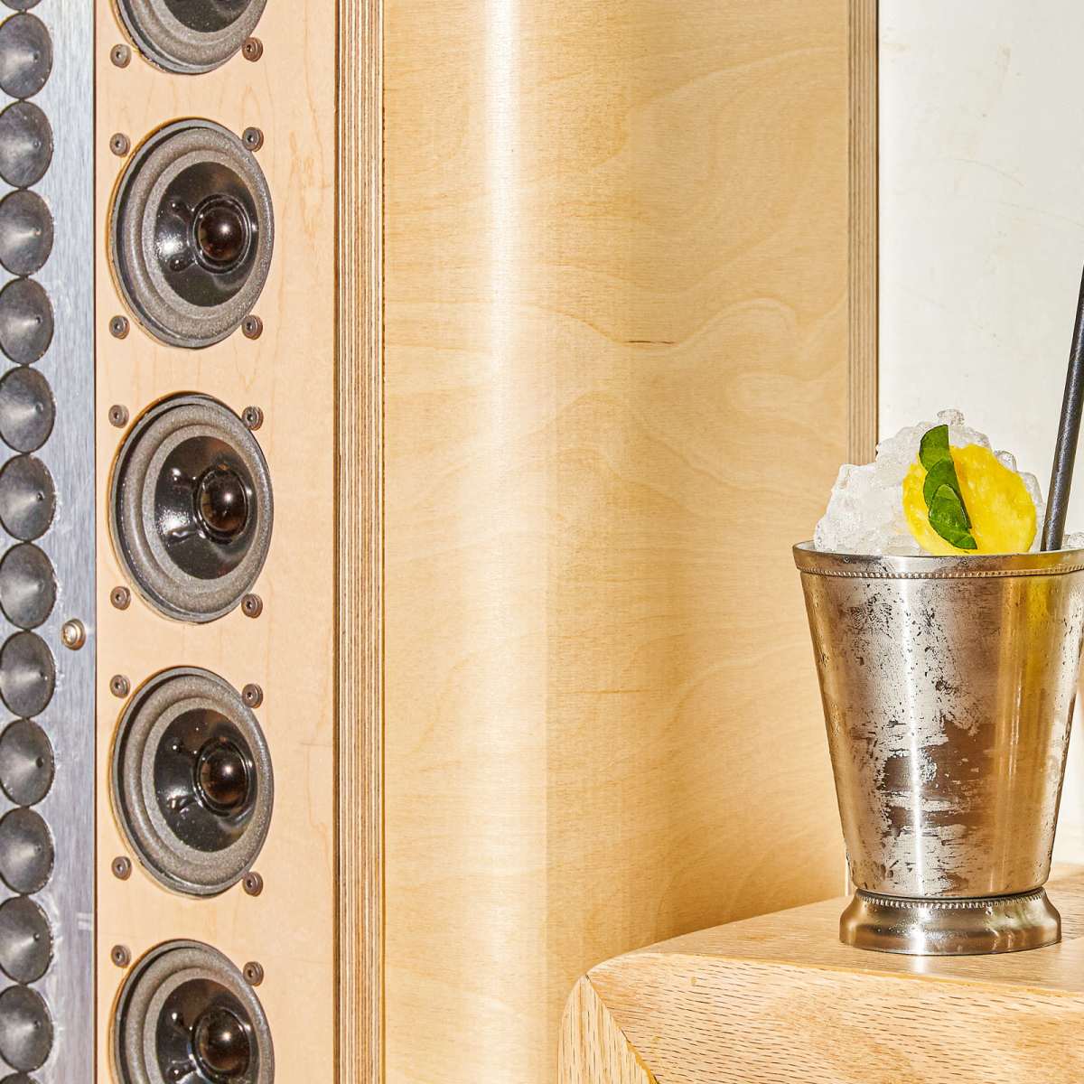 A silver cocktail glass filled with crushed ice, a lime wedge, and a black straw sits next to a bowl of oranges on a wooden surface, with speaker grills in the background.