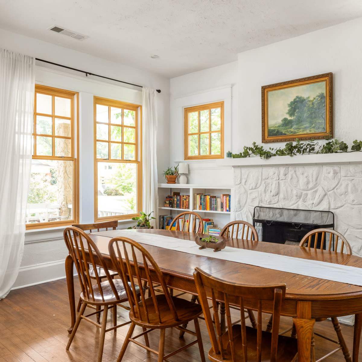 A dining room features a long wooden table surrounded by eight chairs, with large windows and a stone fireplace decorated with a painting and plants.