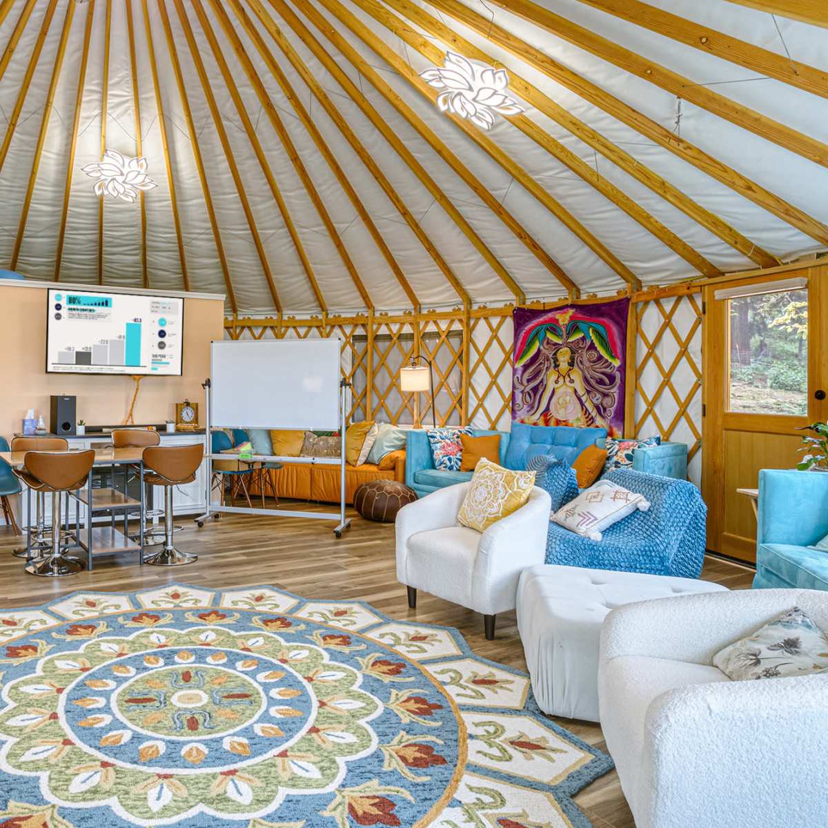 The interior of a yurt, featuring a circular seating area, a decorative rug, and a presentation screen against a backdrop of wooden lattice walls.
