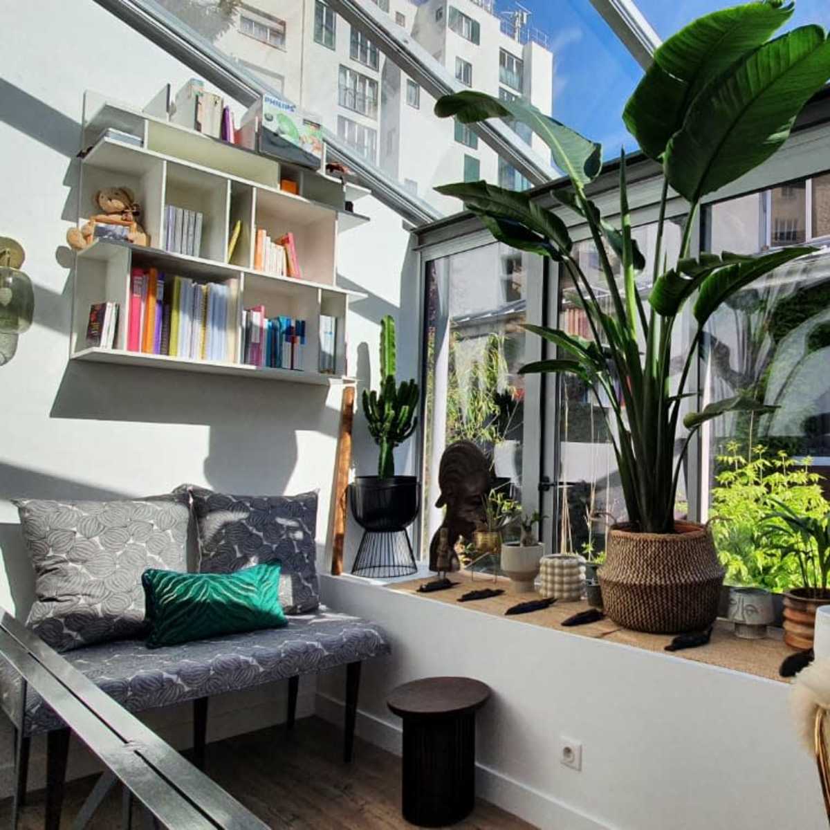 A sunlit indoor garden features a plant-filled window nook with shelves, a bench, and decorative sculptures.