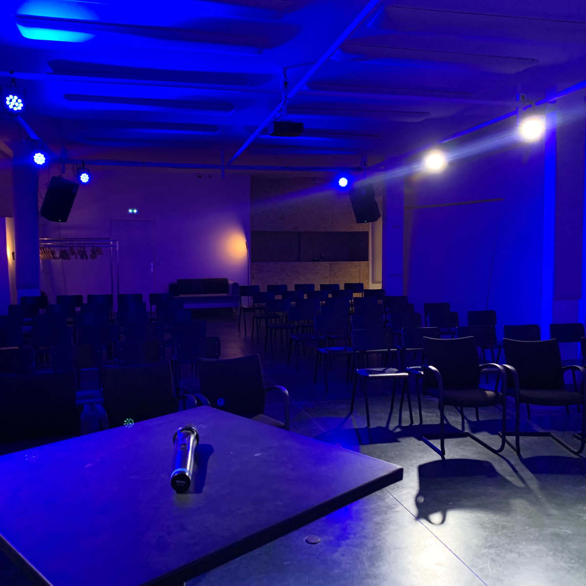 A dimly lit auditorium features rows of black chairs facing a central table under blue stage lighting.