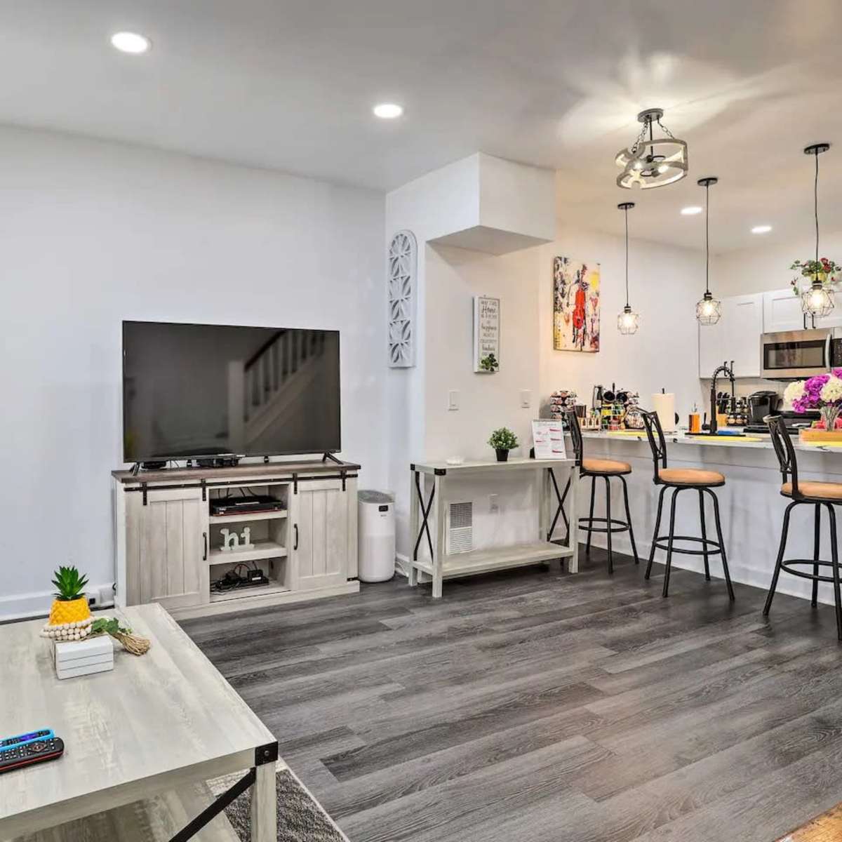 A modern living room features a television unit, a sofa, and an open kitchen with bar stools and pendant lighting.