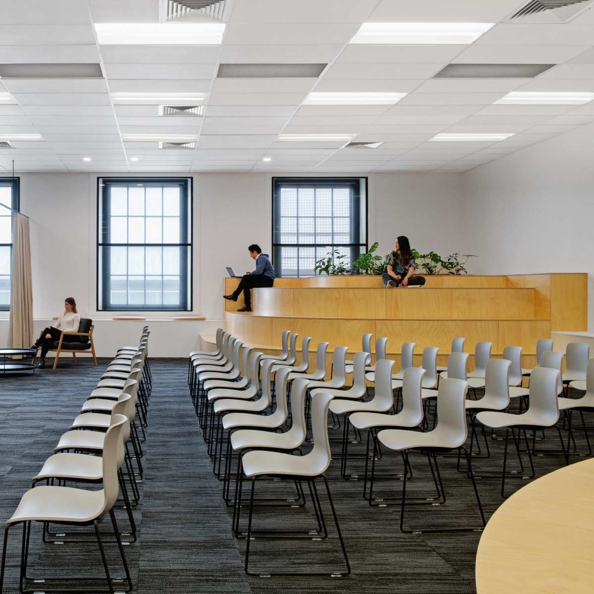 A modern, minimalist office space with rows of chairs facing a large wooden panel that houses a TV, complemented by seating areas and large windows.