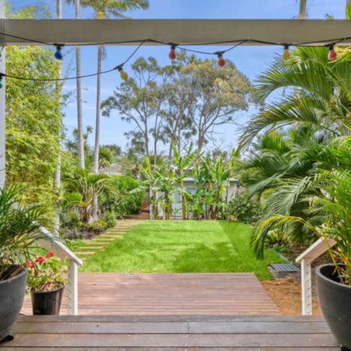 A wooden deck leads to a green lawn surrounded by lush plants and trees.
